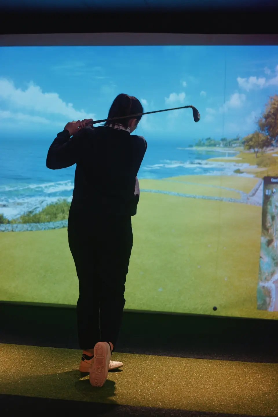 Person playing indoor golf in front of a large screen displaying a golf course scene.