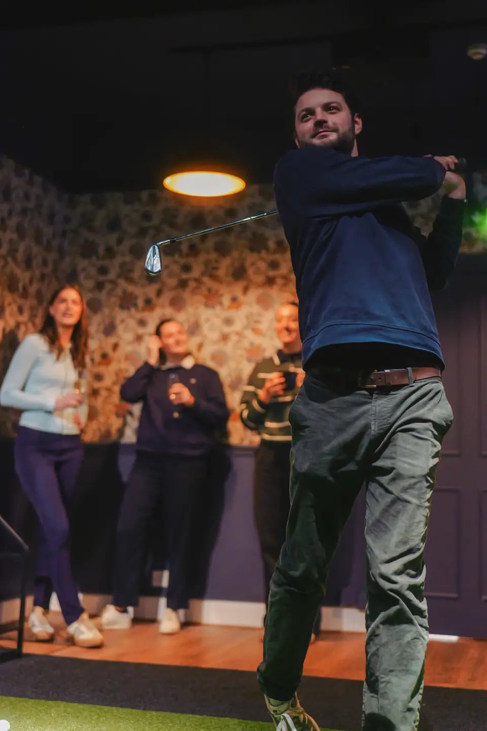A man is swinging a golf club indoors at a gathering, with three women watching and smiling in the background.