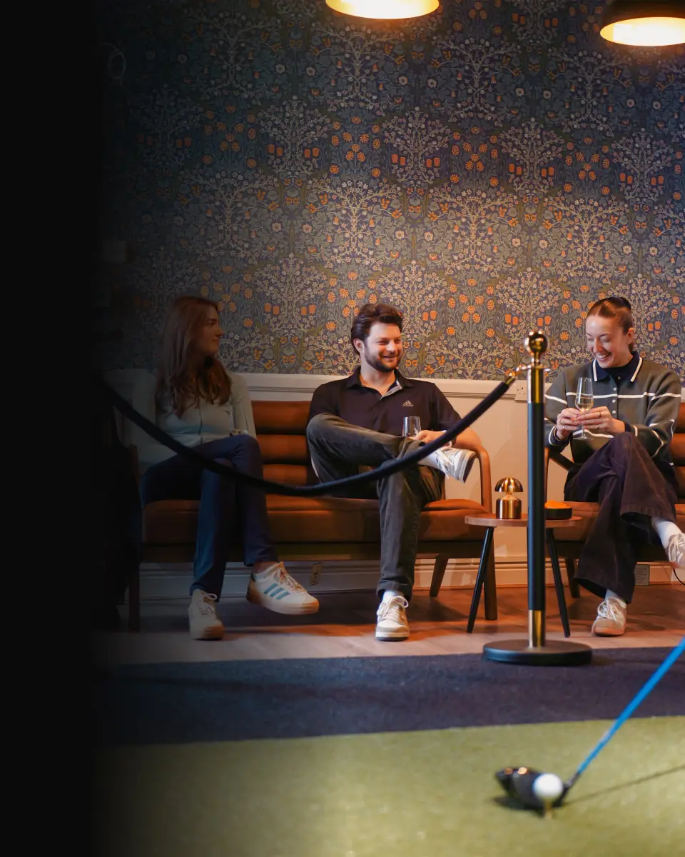 Three people sitting on a bench in a cozy room with floral patterned wallpaper, two women and one man, smiling and engaged in conversation, with a golf putter and ball on the floor in front of them.