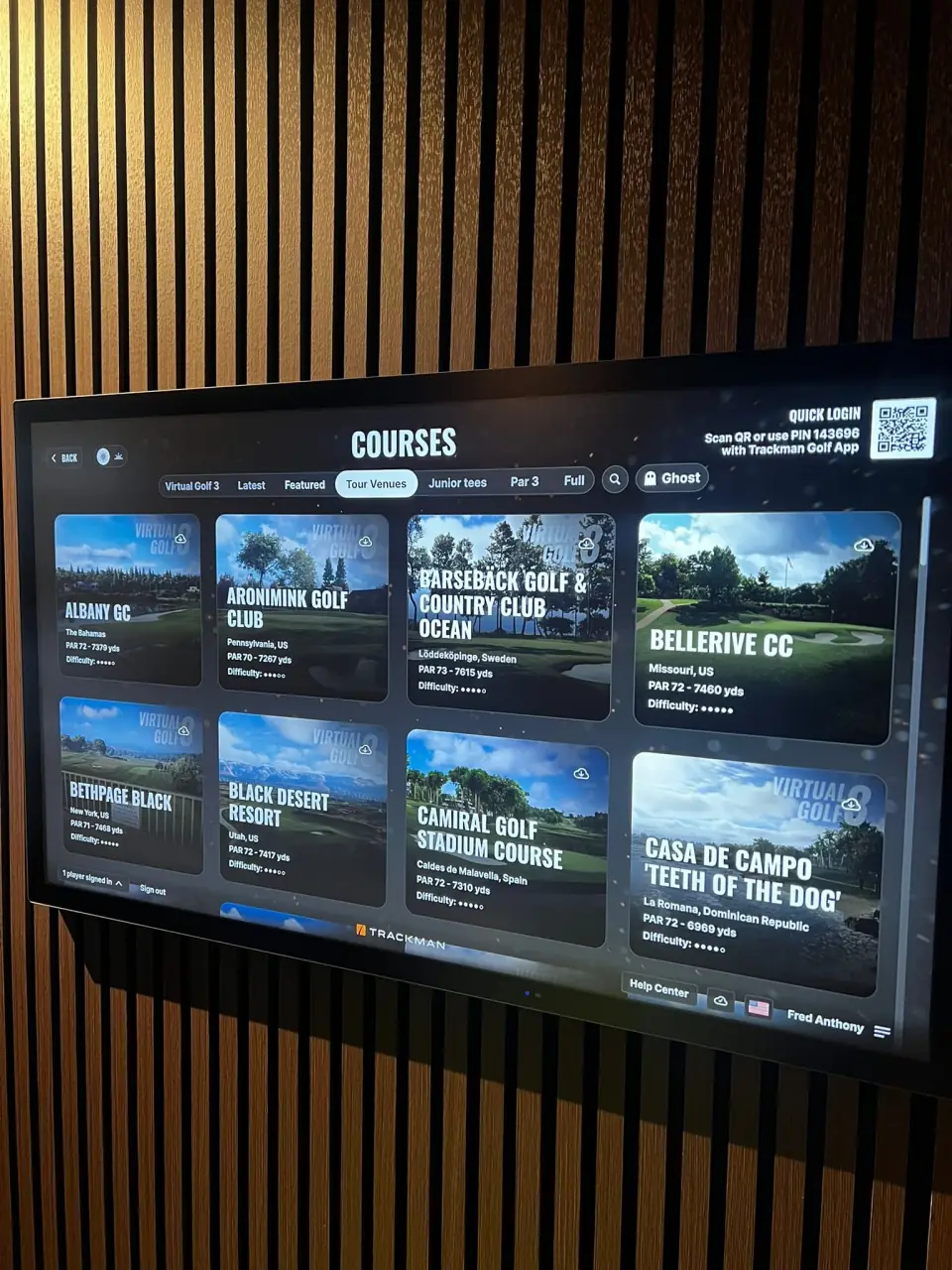 Digital screen displaying list of golf courses at a golf facility, including names, locations, and distances, with categories such as Virtual Golf, Latest, Featured, Tour Venues, Junior Tees, Par 3, and Full, mounted on a wall with vertical wooden paneling.