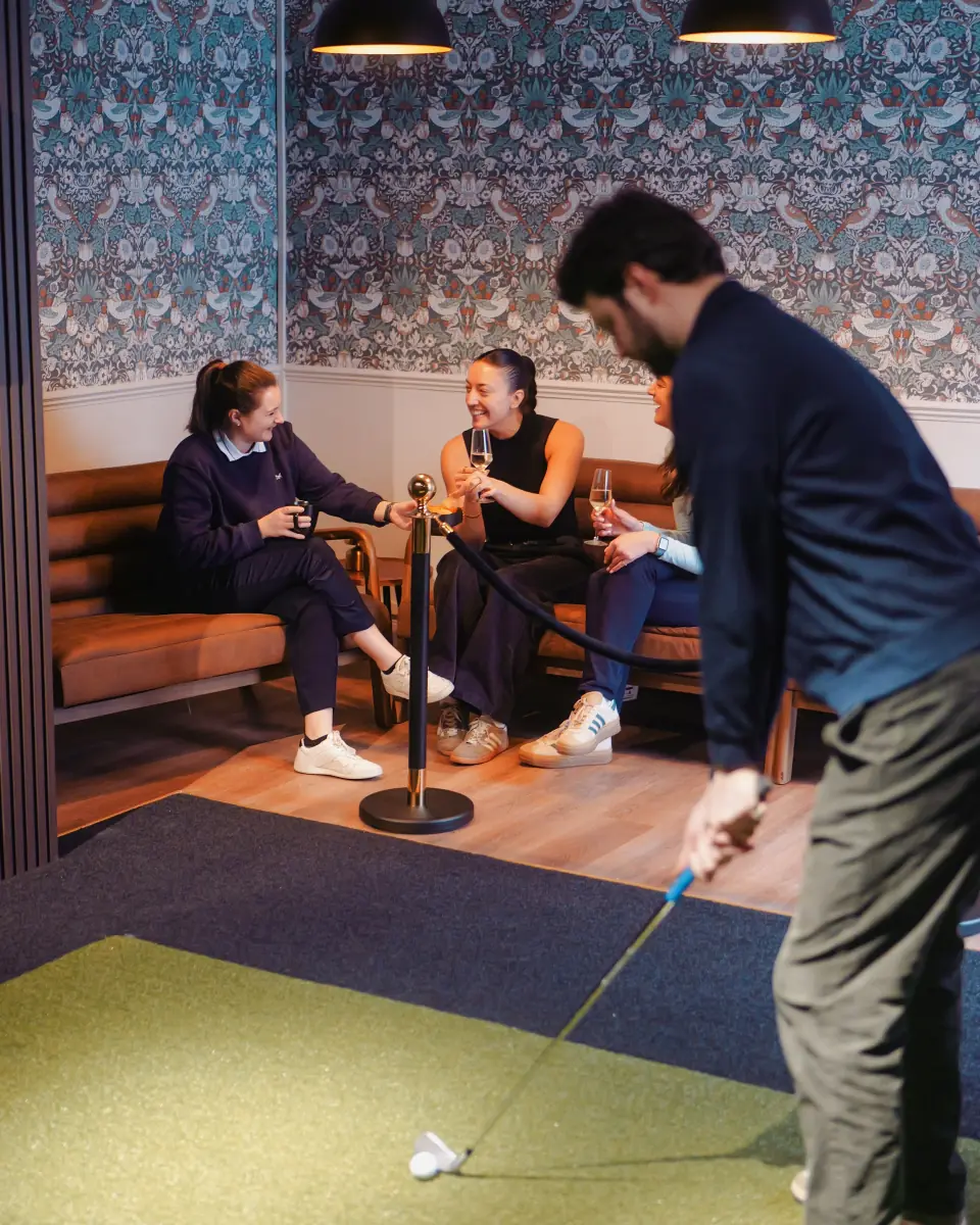 People sitting on a brown cushioned bench in a room with patterned wallpaper, two hanging black lamps, and colorful tiled floor; one person in the foreground is swinging a golf club, partially visible.
