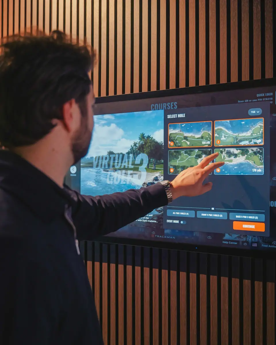 A person in a black jacket with short dark hair is pointing at a screen displaying virtual golf course options, with a wooden wall behind.